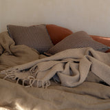 A cozy bedroom scene with a rumpled beige throw blanket and two patterned pillows resting on a bed against a textured beige wall.