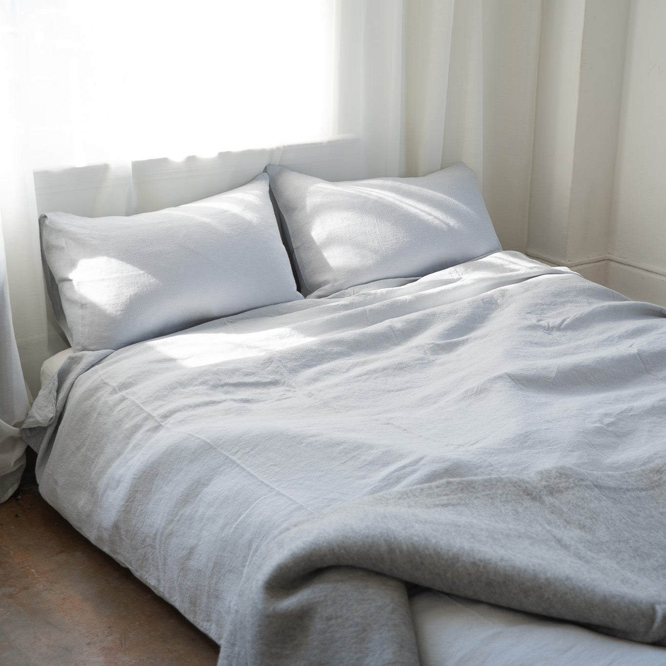 A neatly made bed with white pillows and light gray bedding, positioned in a softly lit bedroom with sheer white curtains and a bright window.