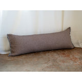 A long rectangular lumbar pillow with a brown and beige houndstooth pattern, resting on a textured concrete floor against a white curtain backdrop.