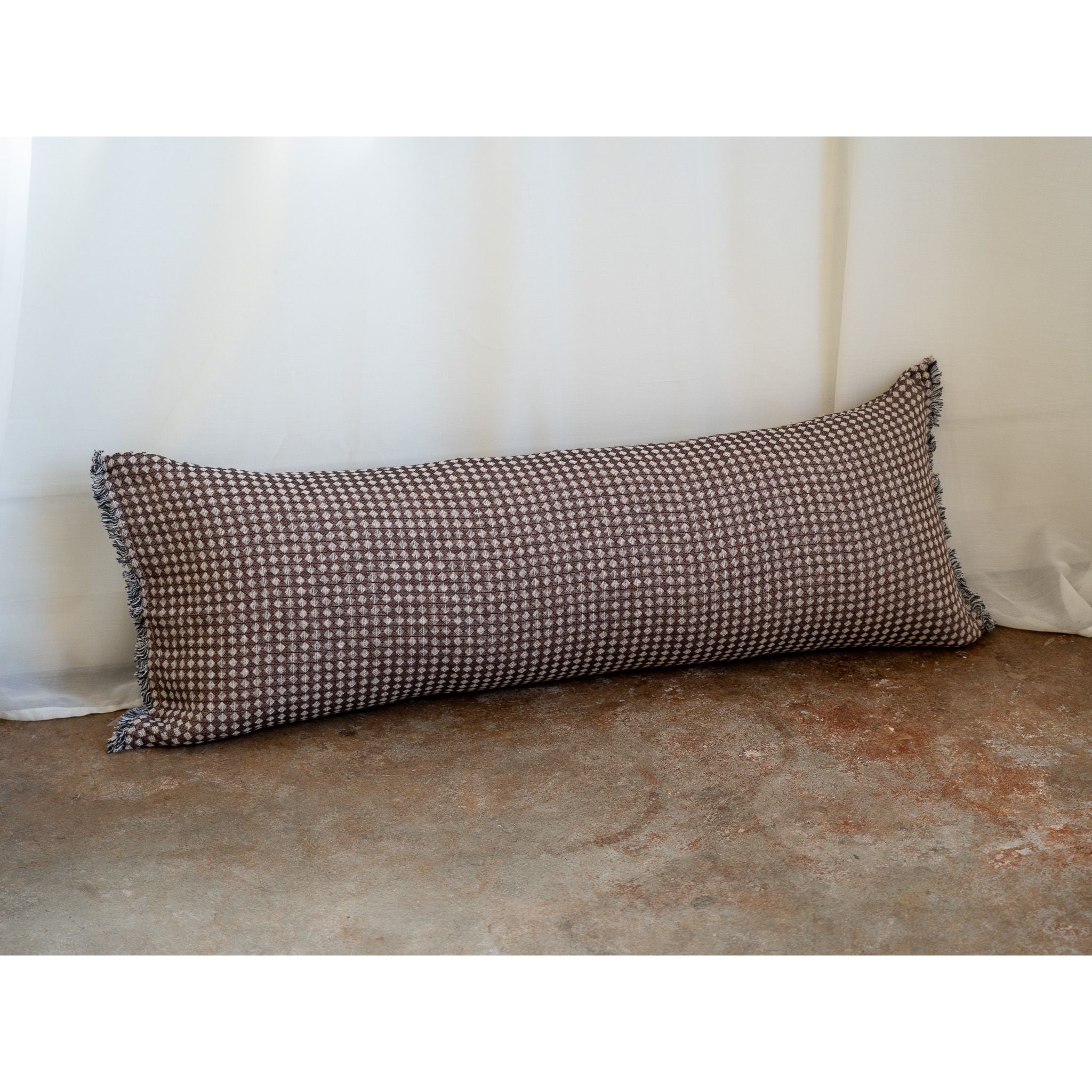 A long rectangular lumbar pillow with a brown and beige houndstooth pattern, resting on a textured concrete floor against a white curtain backdrop.
