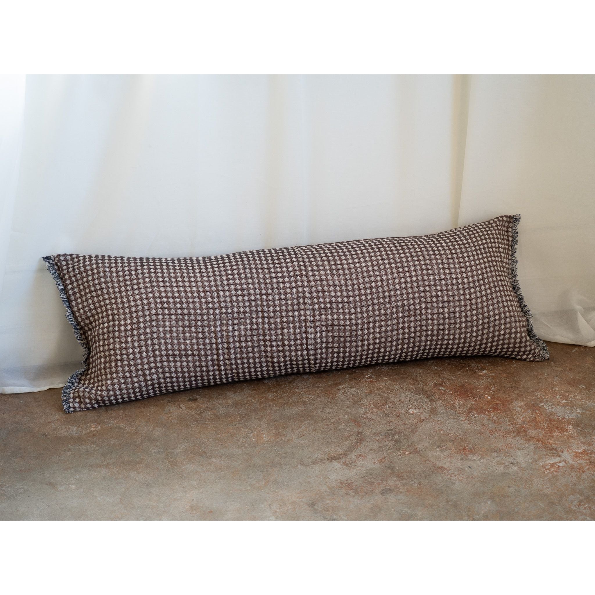 A long rectangular lumbar pillow with a brown and beige houndstooth pattern and fringed edges, resting on a textured concrete floor against a white curtain backdrop.