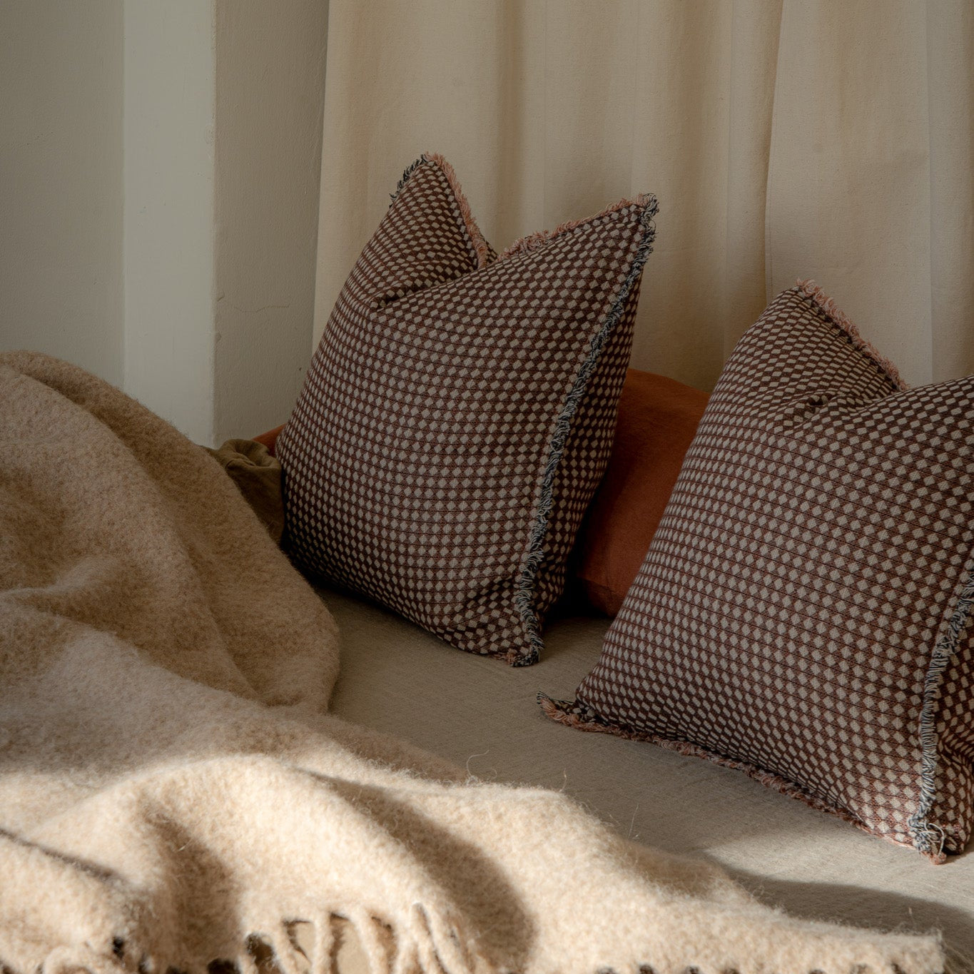 Two brown and beige checkered throw pillows resting on a bed with a cream-colored blanket and a neutral curtain in the background.