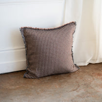 A square throw pillow with a brown and beige houndstooth pattern and fringed edges, resting on a concrete floor against a white paneled wall and curtain.