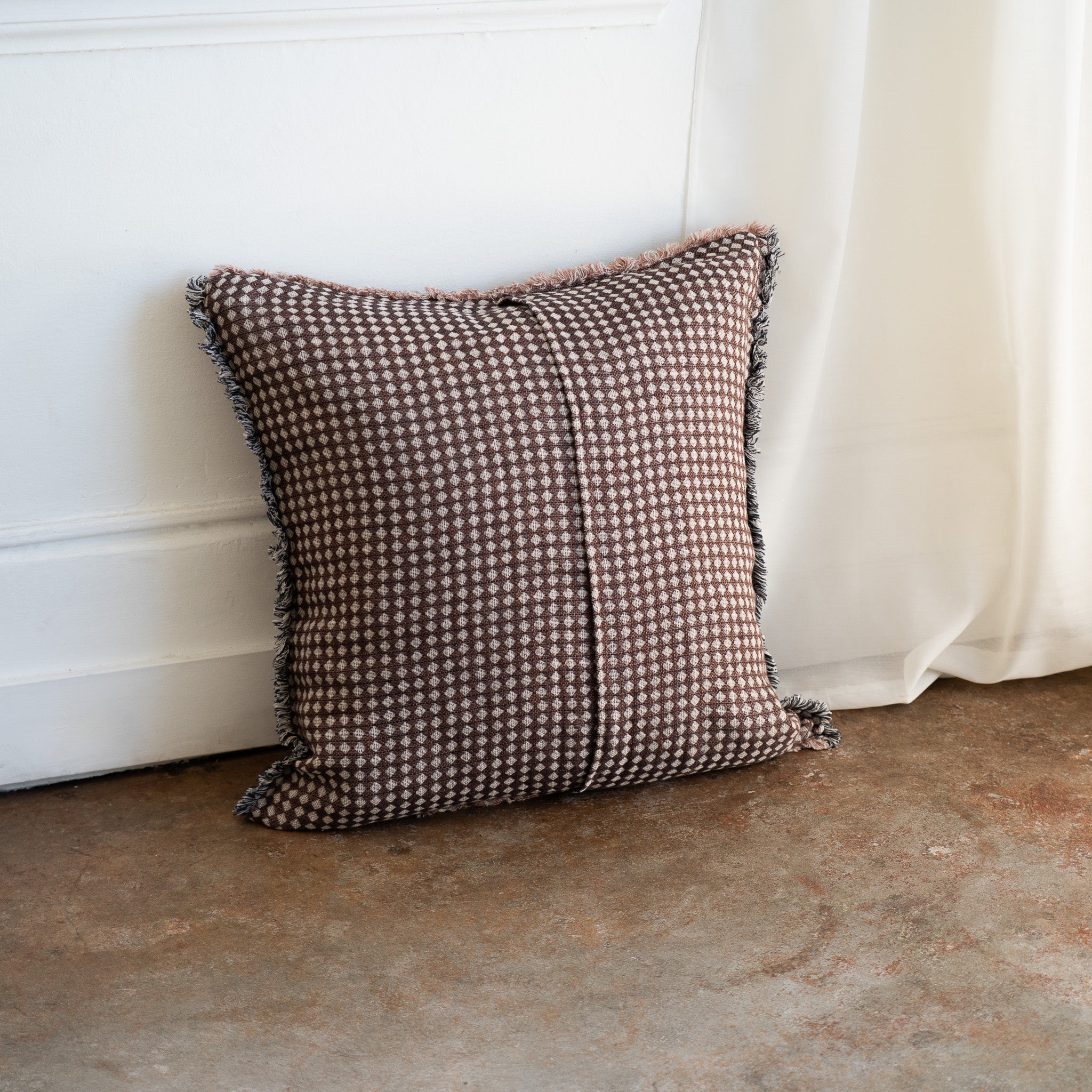 A square throw pillow with a brown and beige houndstooth pattern and fringed edges, leaning against a white wall on a concrete floor next to a white curtain.