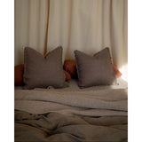 Two square throw pillows with a brown and beige gingham pattern and ruffled edges, resting on a bed with a gray blanket in front of a cream-colored curtain.