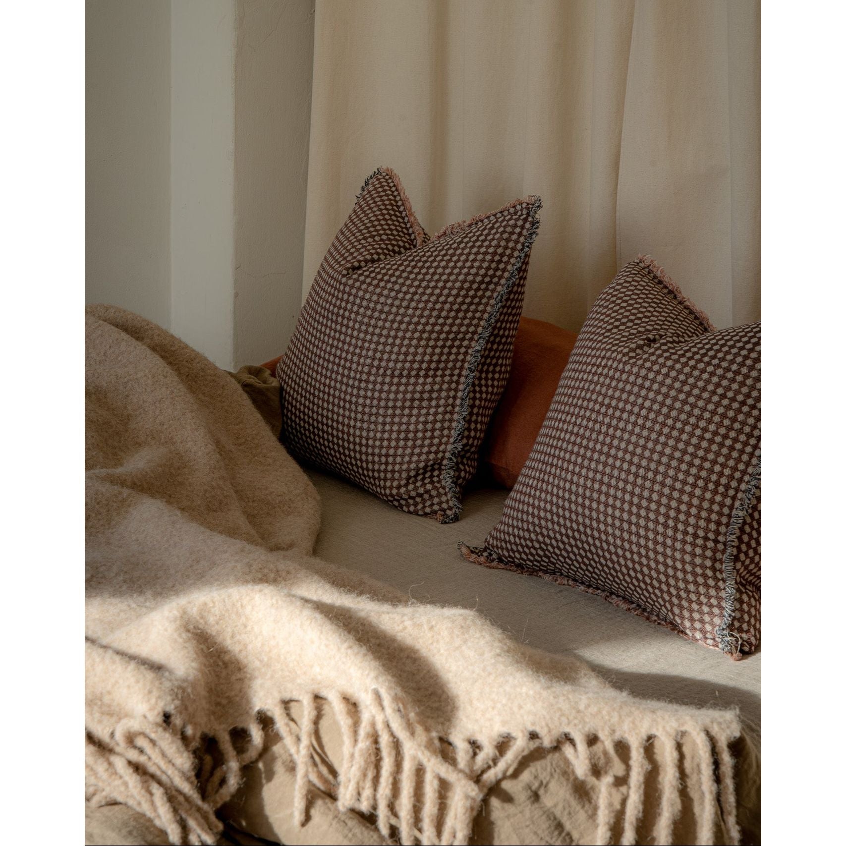 Two brown and beige checkered throw pillows resting on a bed with a cream-colored blanket and a neutral curtain in the background.
