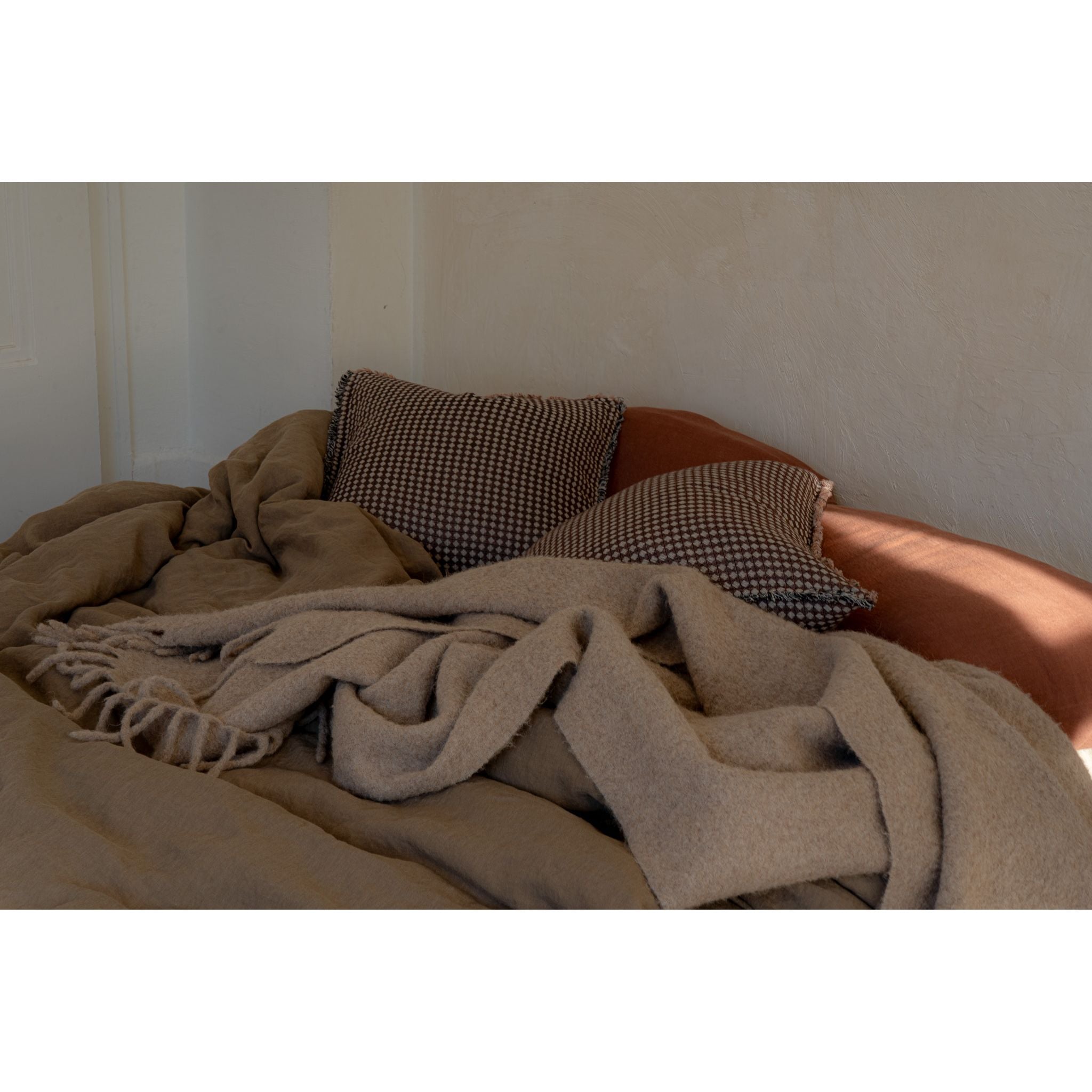 A cozy arrangement of a beige wool blanket draped over a rust-colored upholstered bed, with two brown and black checkered pillows against a textured off-white wall.