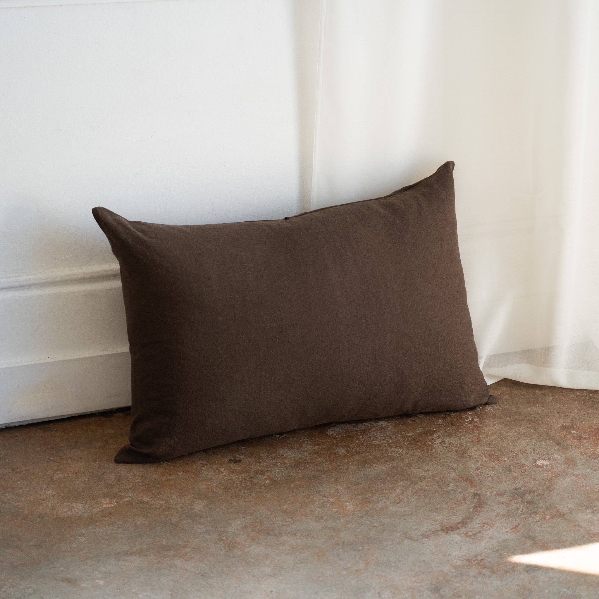A dark brown rectangular throw pillow with a textured linen-like fabric, leaning against a white wall on a rustic concrete floor next to a white curtain.