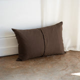 A dark brown rectangular throw pillow with a central seam, resting on a rustic concrete floor against a white wall and curtain backdrop.