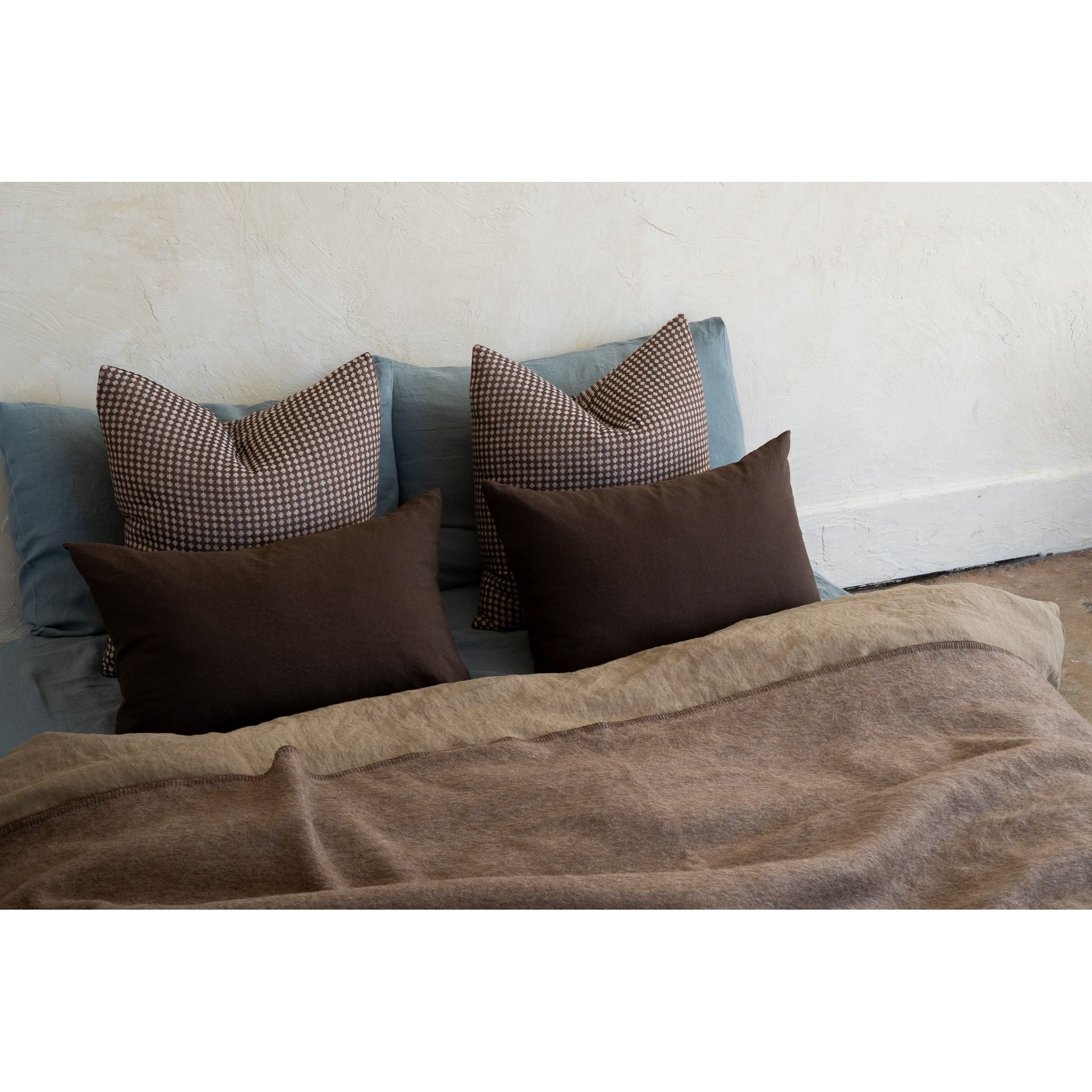 A styled bed setup featuring dark brown throw pillows, a brown and beige checkered pillow, and a blue-gray pillow, layered with a textured brown blanket against a neutral off-white wall.