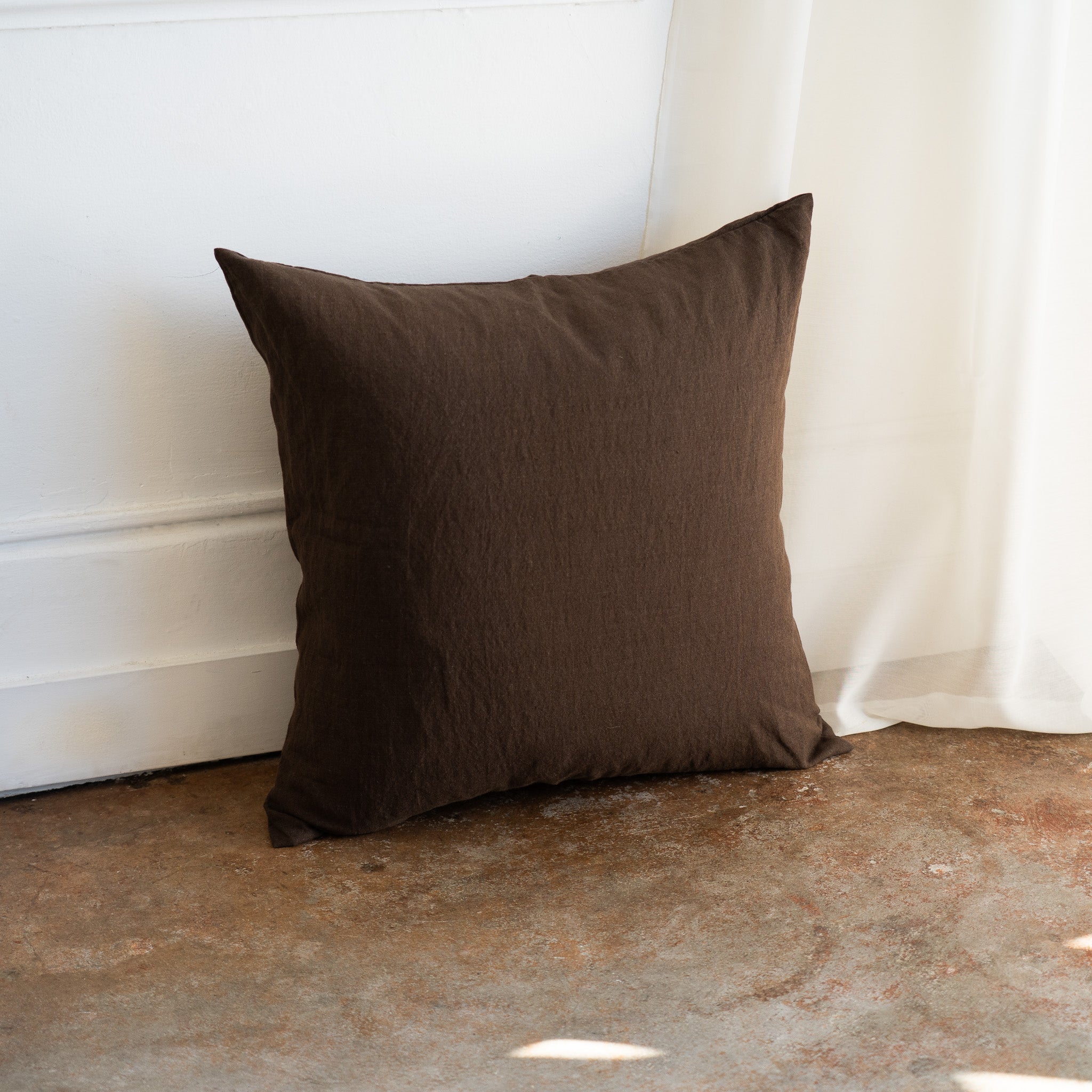 A square dark brown textured throw pillow leaning against a white wall on a rustic concrete floor next to a white curtain.