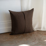 A square brown throw pillow with a central seam, resting on a concrete floor against a white wall and curtain.