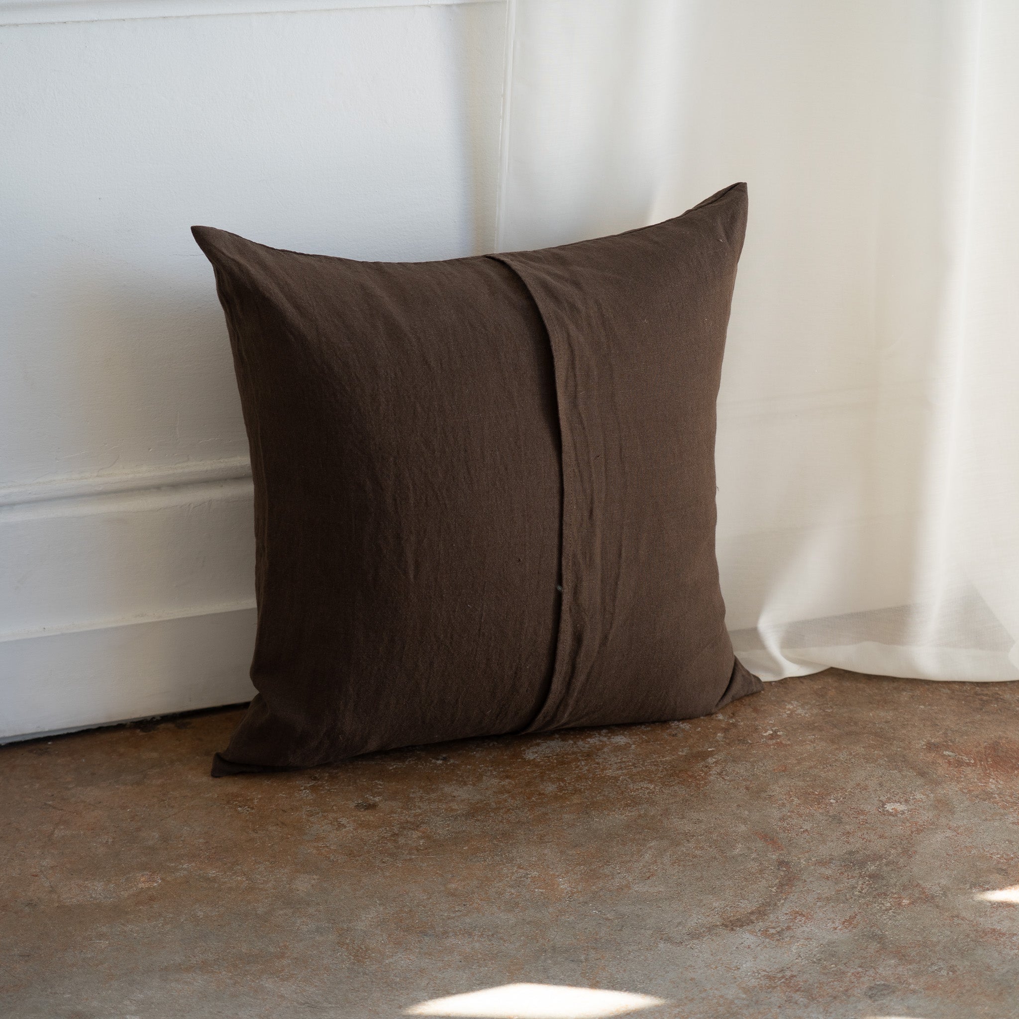 A square brown throw pillow with a central seam, resting on a concrete floor against a white wall and curtain.