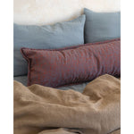 A styled bedroom scene with a rust-red patterned lumbar pillow resting on top of two muted blue-gray pillows, all set against a textured off-white wall and a rumpled beige linen blanket.