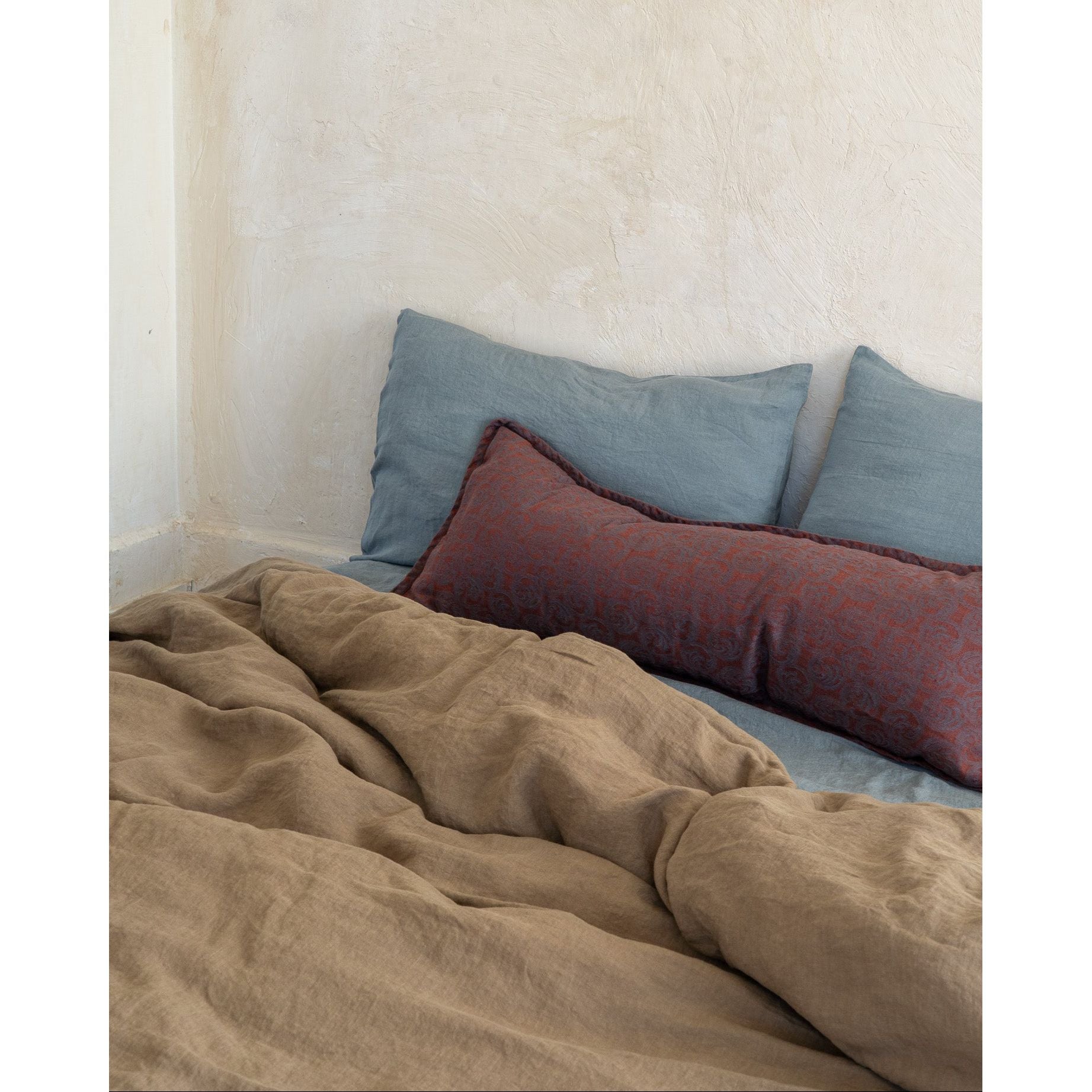 A rustic bedroom scene featuring a textured beige linen duvet and two slate blue pillows, with a rust-red patterned lumbar pillow resting in the foreground against a plastered wall.