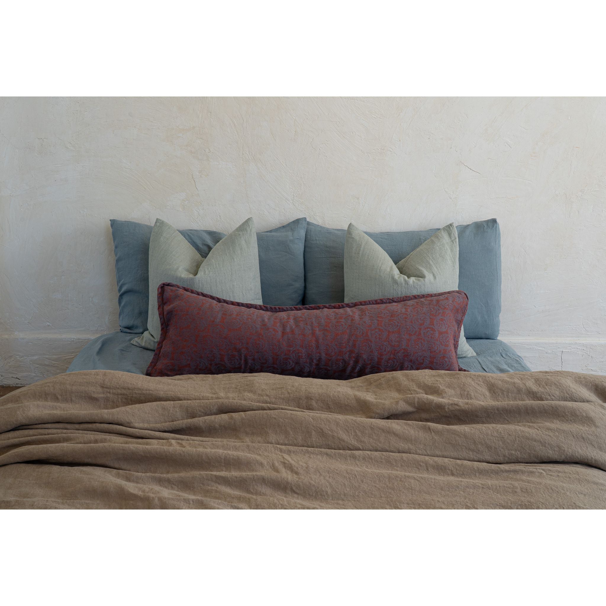 A styled bed setup featuring a slate blue upholstered headboard with multiple pillows, including a prominent textured rust-red lumbar pillow and a beige linen throw blanket in the foreground.
