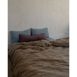 A bed dressed in a textured taupe-brown duvet cover with two slate blue pillows and a burgundy accent pillow, set against a warm beige plaster wall.