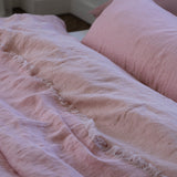 A close-up of light pink crinkled bedding, including a textured duvet cover with frayed edges and a matching pillow, styled on a bed in a softly lit bedroom.