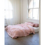 A bed dressed in soft blush pink bedding and matching pillows, placed in a bright minimalist room with white walls, sheer curtains, and a large arched window.