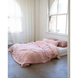 A bed dressed in soft blush pink bedding and matching pillows, placed in a bright minimalist room with white walls, sheer curtains, and a large arched window.
