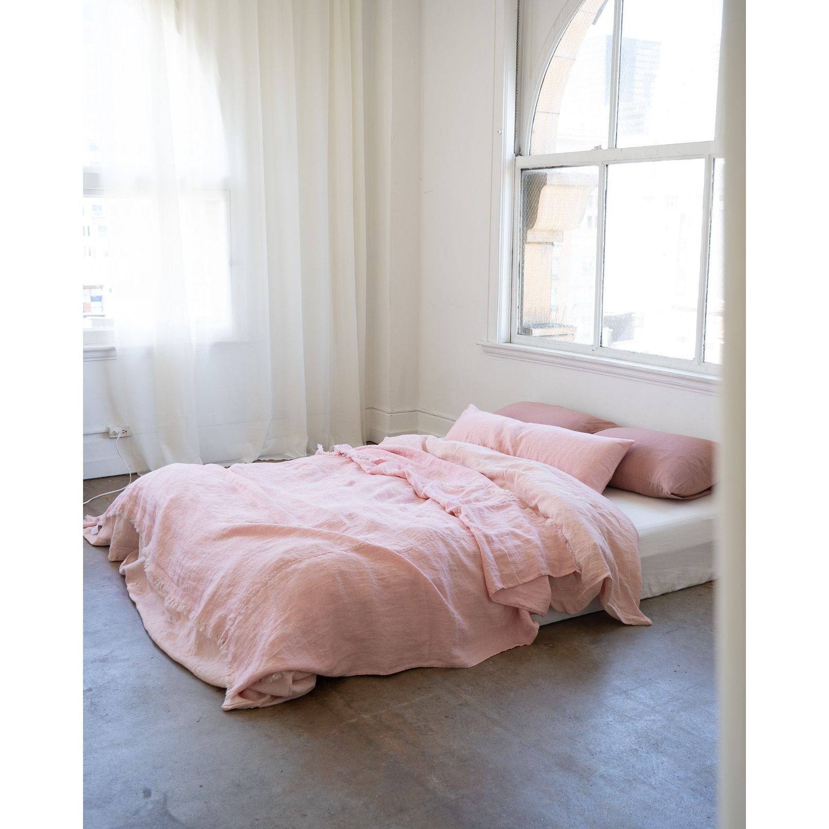 A bed dressed in soft blush pink bedding and matching pillows, placed in a bright minimalist room with white walls, sheer curtains, and a large arched window.
