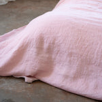 A close-up of a light pink textured linen blanket draped over a bed in a minimalist bedroom setting with a concrete floor and white walls.