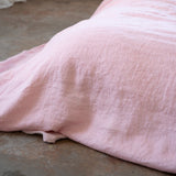 A close-up of a light pink textured linen blanket draped over a bed in a minimalist bedroom setting with a concrete floor and white walls.