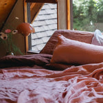 A cozy bedroom scene with a bed dressed in dusty rose and mauve bedding, pillows, and rumpled sheets, set in a rustic wooden loft with a window looking out onto greenery.