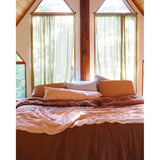 A rustic bedroom scene with a bed dressed in earthy brown and peach bedding, set beneath tall arched windows with sheer olive-green curtains and a wooden ceiling.