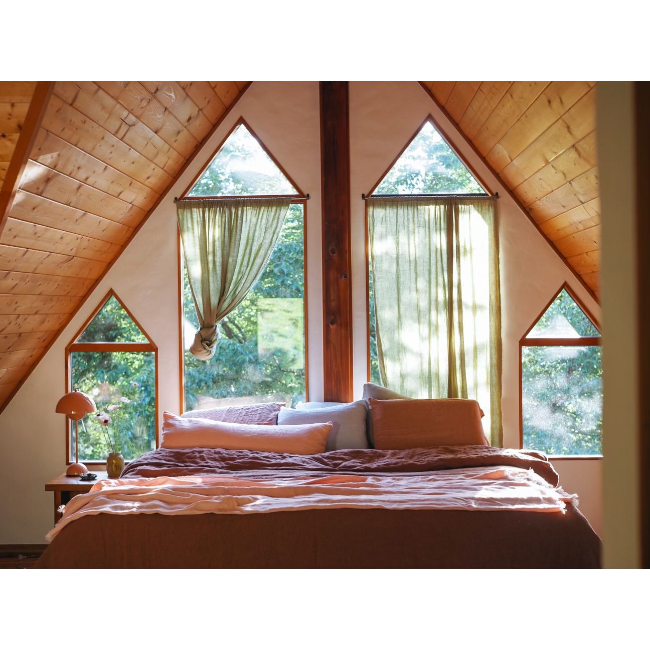 A cozy bedroom with a bed dressed in brown and peach bedding, set under a slanted wooden ceiling with tall arched windows and sheer curtains looking out to green trees.