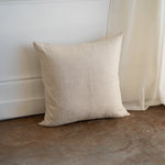 A square beige linen throw pillow with a textured natural fabric, leaning against a white wall on a rustic concrete floor next to a sheer curtain.