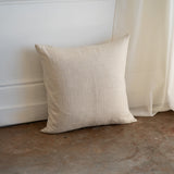 A square beige linen throw pillow with a textured natural fabric, leaning against a white wall on a rustic concrete floor next to a sheer curtain.