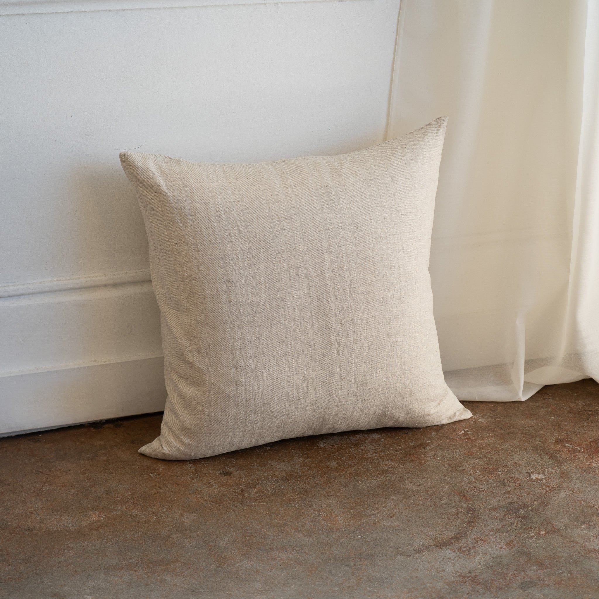 A square beige linen throw pillow with a textured natural fabric, leaning against a white wall on a rustic concrete floor next to a sheer curtain.