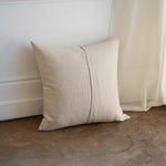 A square beige linen throw pillow with a central vertical seam, leaning against a white wall on a concrete floor next to a sheer curtain.