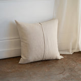 A square beige linen throw pillow with a central vertical seam, leaning against a white wall on a concrete floor next to a sheer curtain.