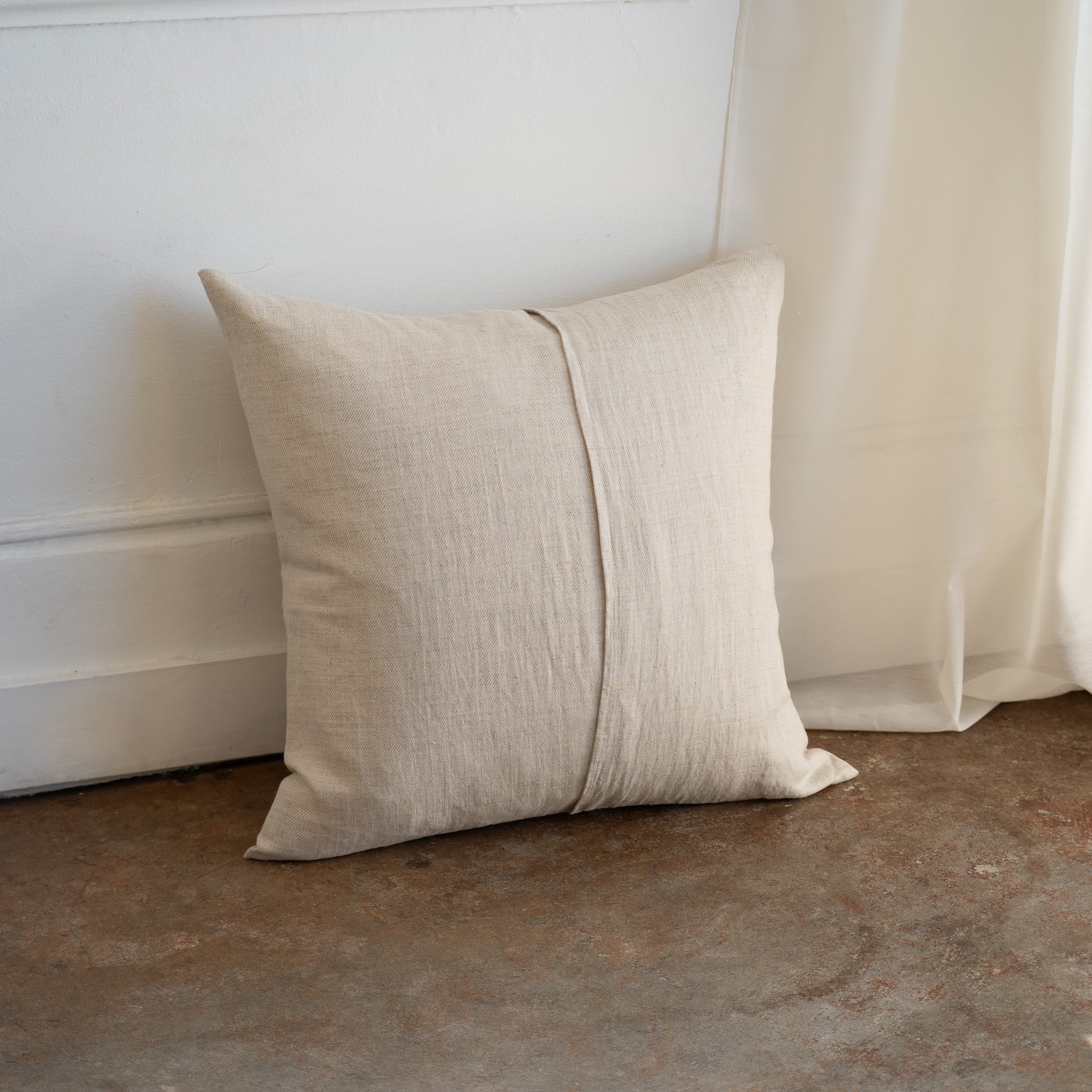 A square beige linen throw pillow with a central vertical seam, leaning against a white wall on a concrete floor next to a sheer curtain.