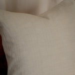 A square off-white linen throw pillow with a textured, natural woven fabric, resting on a neutral gray surface against a soft beige background.