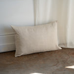 A rectangular beige linen throw pillow with a natural textured weave, leaning against a white wall and sheer curtain on a rustic concrete floor.