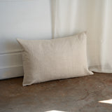 A rectangular beige linen throw pillow with a natural textured weave, leaning against a white wall and sheer curtain on a rustic concrete floor.