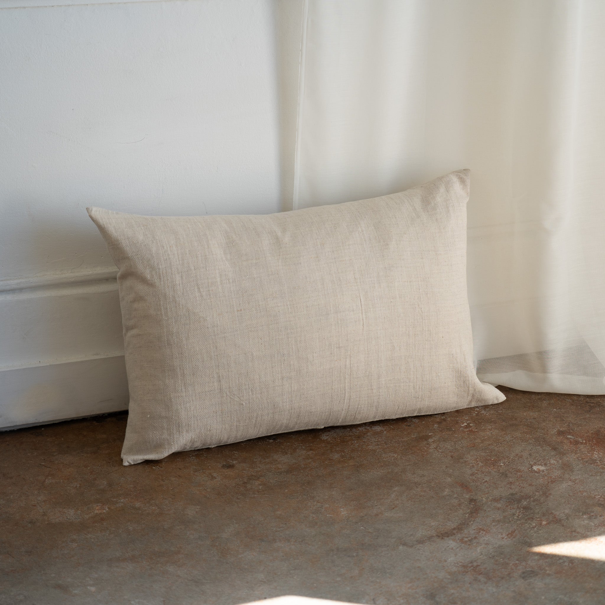 A rectangular beige linen throw pillow with a natural textured weave, leaning against a white wall and sheer curtain on a rustic concrete floor.