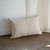 A rectangular beige linen throw pillow with a central vertical seam, resting on a rustic concrete floor against a white paneled wall and sheer curtain.
