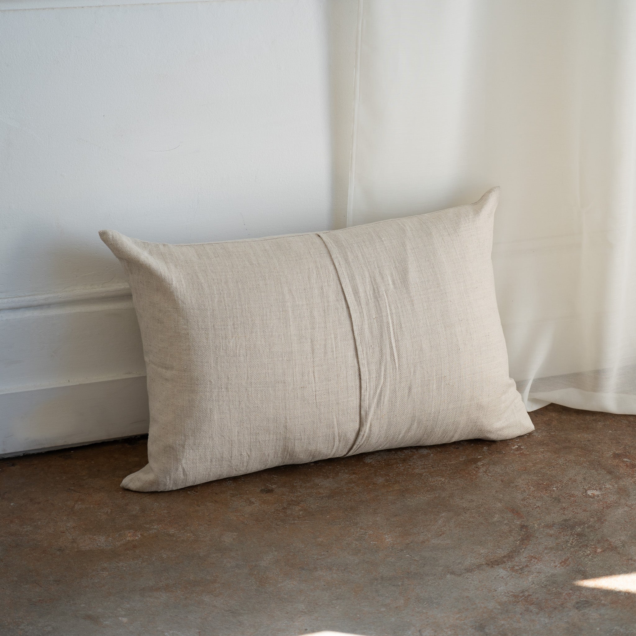 A rectangular beige linen throw pillow with a central vertical seam, resting on a rustic concrete floor against a white paneled wall and sheer curtain.