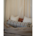 A cozy bed setup with a textured geometric plaid lumbar pillow in front of two white pillows and a rust-colored headboard, set against a backdrop of cream-colored curtains.