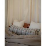 A cozy bed setup with a textured geometric plaid lumbar pillow in front of two white pillows and a rust-colored headboard, set against a backdrop of cream-colored curtains.