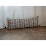 A long rectangular lumbar pillow with a textured geometric plaid pattern in beige, brown, and muted red, resting on a concrete floor in front of white draped curtains.