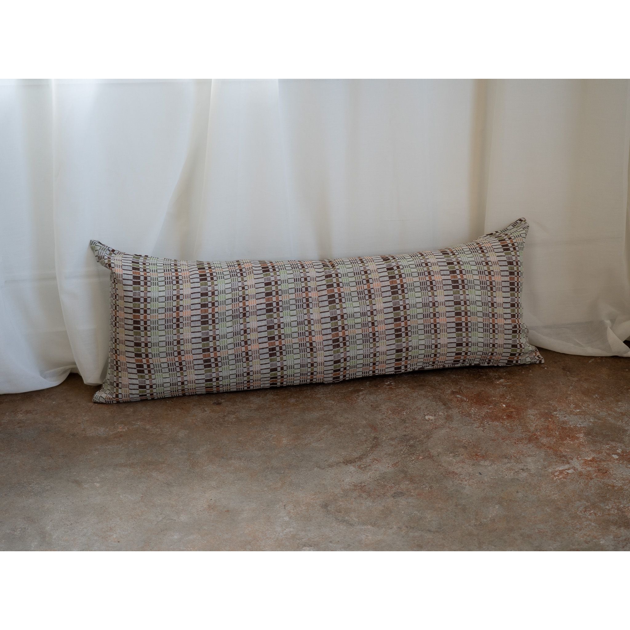 A long rectangular lumbar pillow with a textured geometric plaid pattern in beige, brown, and muted red, resting on a concrete floor in front of white draped curtains.