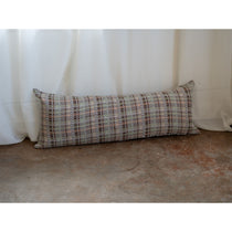 A long rectangular lumbar pillow with a textured geometric plaid pattern in beige, brown, and muted red, resting on a concrete floor in front of white draped curtains.