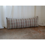A long rectangular lumbar pillow with a textured plaid pattern in beige, brown, and gray, resting against a white curtain on a concrete floor.