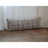 A long rectangular lumbar pillow with a textured plaid pattern in beige, brown, and gray, resting against a white curtain on a concrete floor.