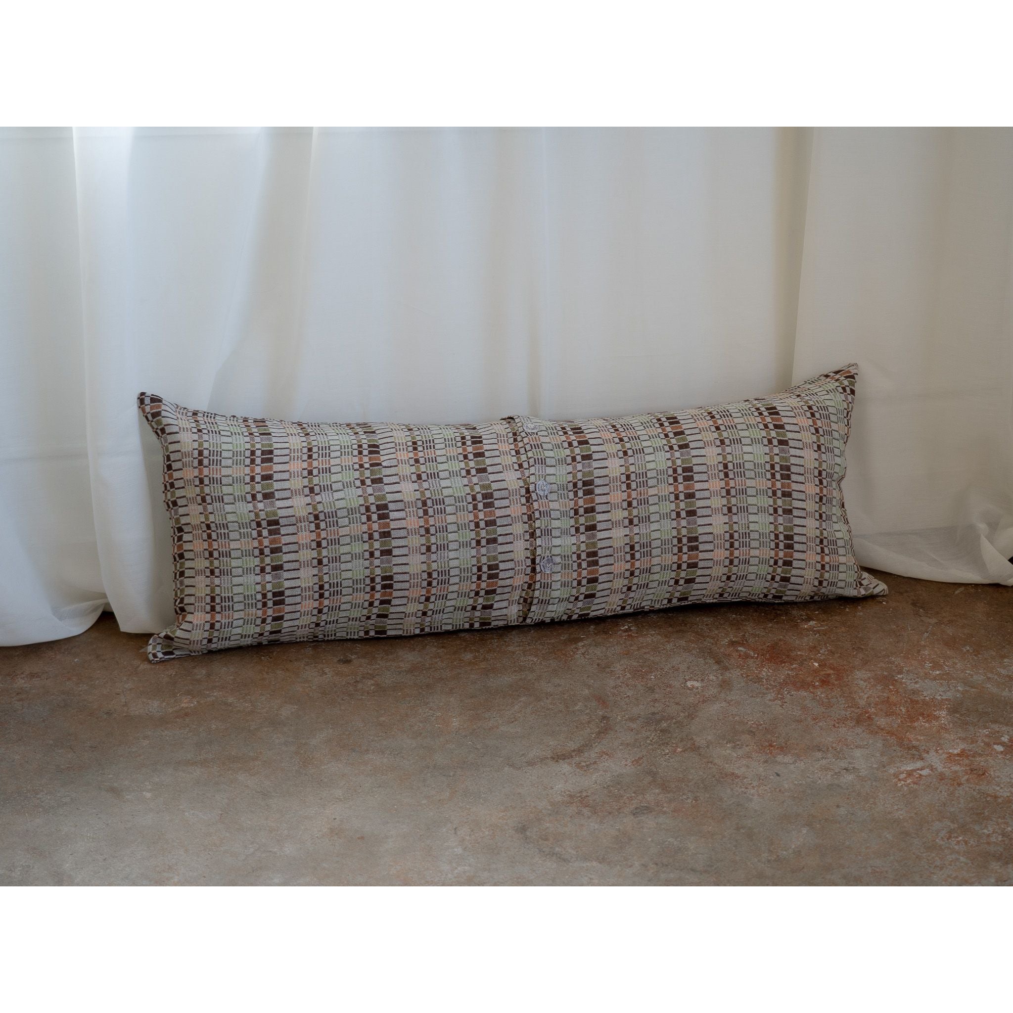 A long rectangular lumbar pillow with a textured plaid pattern in beige, brown, and gray, resting against a white curtain on a concrete floor.